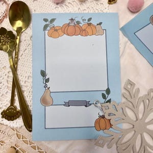 Op de afbeelding: Een lichtblauwe kaart met een pompoenillustratie bovenaan en een banner onderaan. De kaart is omgeven door decoratieve items, waaronder een gouden lepel, roze pompons en een sneeuwvlokornament.