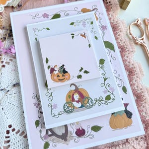 Könnte beinhalten: Ein Set aus gestapeltem Briefpapier in Hellrosa mit Illustrationen von Kürbissen, Ranken und kleinen Blumen. Der obere quadratische Zettel zeigt zwei kleine Figuren in einem Kürbis, während die größere rechteckige Karte eine Kürbiskutsche zeigt.