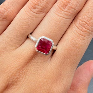 May include: A silver ring featuring a large, rectangular, deep red gemstone. The gemstone is set in a bezel setting, and the ring band is a simple, polished silver. The ring is shown on a person's finger.
