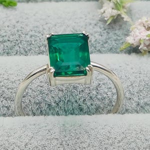 Puede incluir: Un anillo de tono plateado con una gran piedra preciosa verde de corte esmeralda rectangular, sujeta por cuatro garras. La banda es fina y lisa, conectándose a la montura que eleva la vibrante piedra verde sobre la base del anillo.