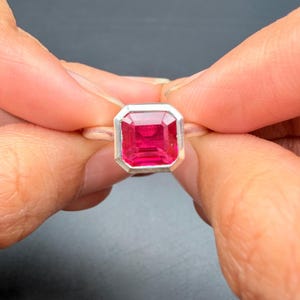 May include: A silver ring with a large, rectangular, bright red gemstone. The gemstone is set in a bezel setting. The ring is held between two hands against a dark background. The ring is a piece of jewellery.