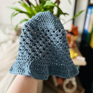 Könnte beinhalten: Ein blauer gehäkelter Fischerhut mit ausgestellter Krempe. Der Hut ist aus einem strukturierten, offen gewebten Stoff gefertigt. Der Hut hat eine hellblaue Farbe und scheint handgefertigt zu sein. Der Hut ist ein Sommeraccessoire.