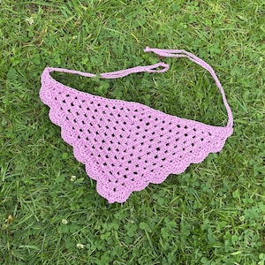 Könnte beinhalten: Ein handgefertigtes, helllila gehäkeltes Bandana liegt auf grünem Gras. Das Bandana hat einen gewellten Rand und Bindebänder im Nacken. Das offene Webmuster ist sichtbar.