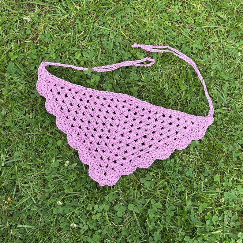 Może przedstawiać: Ręcznie robiona, jasnofioletowa chusta bandana z szydełka leży na zielonej trawie. Bandana ma falisty brzeg i wiązania z tyłu szyi. Widoczny jest wz&oacute;r z otwartym splotem.
