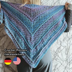 Puede incluir: Un chal de crochet hecho a mano, de tejido abierto, en tonos de azul, morado y verde azulado. El chal triangular presenta un diseño de chevrón y un borde festoneado. El texto "Crochet Pattern Häkelanleitung" se muestra con las banderas de Alemania y Estados Unidos.