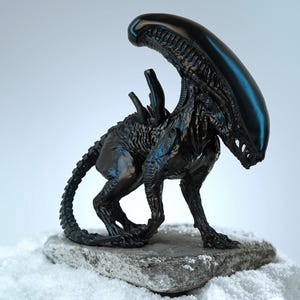 Può includere: Una statuetta nera di Xenomorfo con testa lucida e corpo strutturato, in piedi su una pietra. La creatura ha una lunga coda segmentata e posa su uno sfondo innevato. I riflessi blu accentuano il design.