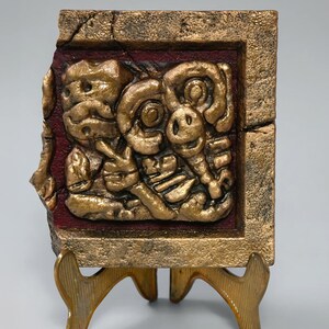 Op de afbeelding: Een vierkante, getextureerde goudkleurige reliëfplaat met een centrale gebeeldhouwde figuur met een opgeheven hand, tegen een dieprode achtergrond in een dikke, verweerde gouden lijst, geplaatst op een kleine driepotige gouden metalen standaard.