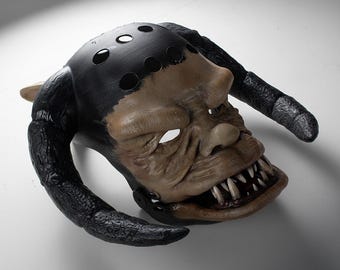 Könnte beinhalten: Eine Kreaturenmaske mit einem gebräunten, faltigen Gesicht, einem breiten Mund, spitzen Zähnen und einer markanten Nase. Die Oberseite ist schwarz mit kreisförmigen Aussparungen und zwei großen, texturierten schwarzen Hörnern.