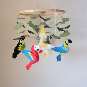 May include: A colorful felt bird mobile featuring a wooden hoop and hanging felt birds. The mobile includes a toucan, a red macaw, a yellow and white cockatoo, and a blue and yellow parrot, along with green felt leaves.