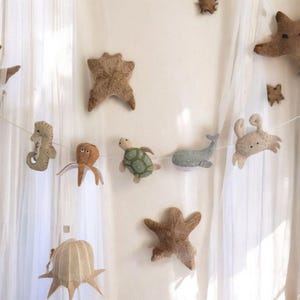 Puede incluir: Guirnalda de criaturas marinas de fieltro, incluyendo un caballito de mar, un pulpo, una tortuga, una ballena y un cangrejo. Las criaturas son en tonos beige, naranja, verde y gris, con estrellas de mar y otras criaturas marinas dispersas.
