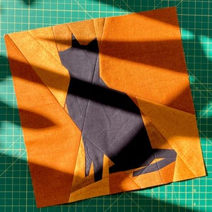 May include: A square, orange and black fabric art piece featuring a silhouette of a cat. The cat is black, and the background is orange with geometric shapes. The piece is likely handmade and suitable for wall decor.