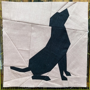 May include: A fabric patchwork block featuring a geometric design of a seated wolf. The wolf is rendered in dark blue fabric against a background of light pink fabric. The block is square and has a ruler along the edges.