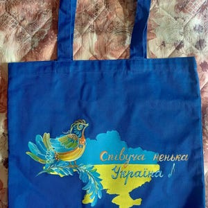 May include: A blue tote bag with a bird design and the text "Singing Ukraine". The bird is multi-colored, and the map outline is yellow and blue. The bag has blue handles.