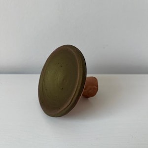 Puede incluir: Un tapón de botella de cerámica con una parte superior redonda de color verde oliva y una pequeña base redondeada de color marrón rojizo. El tapón está diseñado para encajar en la abertura de una botella. El objeto está sobre un fondo blanco.