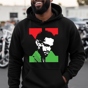 May include: Black hoodie featuring a graphic portrait of a man with glasses, set against a red and green backdrop. The hoodie has a drawstring hood and a front pocket. The person in the image is wearing a black cap and sunglasses.