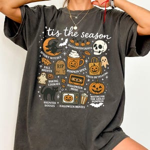 May include: Dark gray t-shirt with a Halloween-themed graphic. The design includes a witch on a crescent moon, pumpkins, a skull, and the phrase "'tis the season." Additional elements include ghosts, a black cat, and the words "spooky witches" and "Halloween movies."