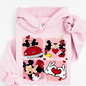 May include: A pale pink hoodie with a graphic of Mickey and Minnie Mouse in romantic scenes. The design includes red hearts and a red car, with Mickey and Minnie kissing and holding hands. The hoodie has a drawstring hood and long sleeves.