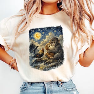 May include: Cream-colored t-shirt featuring a whimsical illustration of two monkeys embracing under a starry night sky with a full moon. A rooster and a bird are also depicted. The artwork is framed by a dark blue border.