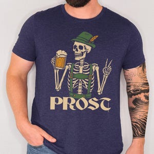 May include: Navy blue t-shirt featuring a skeleton wearing a green hat and suspenders, holding a beer stein. The word "PROST" is printed in a vintage font below the skeleton. The shirt has a crew neck.