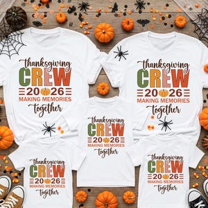May include: Five white t-shirts featuring the text "thanksgiving CREW 2026 MAKING MEMORIES together" with a pumpkin graphic. The shirts are arranged on a wooden surface decorated with small pumpkins, spider webs, and plastic spiders.