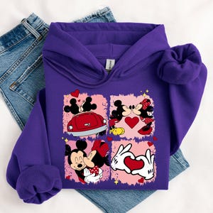 May include: A purple hoodie with a graphic of Mickey and Minnie Mouse in four romantic scenes. The design includes a red car, hearts, and the couple kissing. The hoodie is folded on top of a pair of blue jeans.