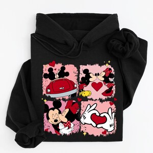May include: Black hoodie with a vibrant graphic of Mickey and Minnie Mouse in romantic scenes. The design incorporates hearts, a red car, and a heart-shaped hand gesture, set against a pink backdrop.