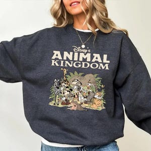 May include: Charcoal grey sweatshirt with the text "Disney's Animal Kingdom" in cream. The graphic depicts cartoon animals including a zebra, giraffe, and tiger, set against green foliage. A casual, comfortable top.