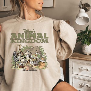 May include: Beige sweatshirt with the text "Disney's Animal Kingdom" in green. The graphic shows cartoon animals like a giraffe, zebra, elephant, and tiger in a jungle scene. The sweatshirt has long sleeves.