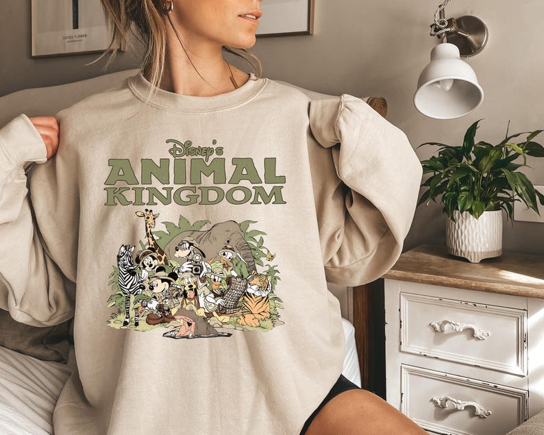 May include: Beige sweatshirt with the text "Disney's Animal Kingdom" in green. The graphic shows cartoon animals like a giraffe, zebra, elephant, and tiger in a jungle scene. The sweatshirt has long sleeves.