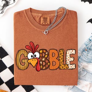 May include: A rust-colored crewneck t-shirt featuring a graphic of a cartoon turkey head replacing the letter O in the word GOBBLE. The letters have various patterns including wood grain, autumn leaves, polka dots, and leopard print.