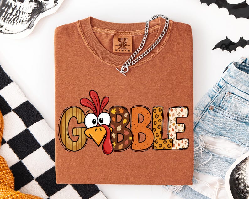 May include: A rust-colored crewneck t-shirt featuring a graphic of a cartoon turkey head replacing the letter O in the word GOBBLE. The letters have various patterns including wood grain, autumn leaves, polka dots, and leopard print.
