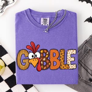May include: A purple t-shirt featuring the word GOBBLE in large, multi-patterned letters. The letter O is replaced by a cartoon turkey face with a red wattle and orange beak. The other letters feature wood grain, autumn leaves, and leopard print patterns.