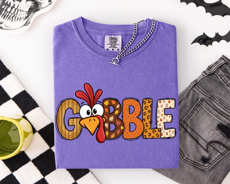 May include: A purple t-shirt featuring the word GOBBLE in large, multi-patterned letters. The letter O is replaced by a cartoon turkey face with a red wattle and orange beak. The other letters feature wood grain, autumn leaves, and leopard print patterns.
