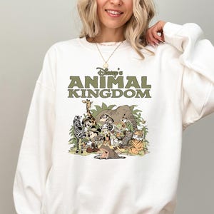 May include: White crew neck sweatshirt with the text "Disney's Animal Kingdom" in green. The graphic features cartoon animals including a zebra, giraffe, elephant, and tiger. The sweatshirt is a casual style.