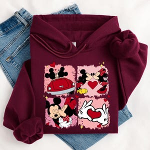 May include: Burgundy hoodie with a graphic of Mickey and Minnie Mouse in romantic scenes. The design includes a red car, hearts, and the couple kissing. The hoodie is displayed on a pair of blue jeans.