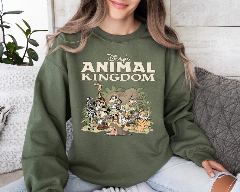 May include: An olive green sweatshirt with the text "Disney's Animal Kingdom" and a graphic of cartoon animals. The graphic includes a giraffe, zebra, elephant, and tiger. The sweatshirt has long sleeves and a crew neck, perfect for casual wear.