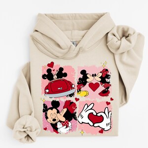 May include: Beige hoodie with a four-panel design showcasing Mickey and Minnie Mouse illustrations. The panels depict romantic scenes with hearts and a red car. The design includes yellow stars and a pink background.