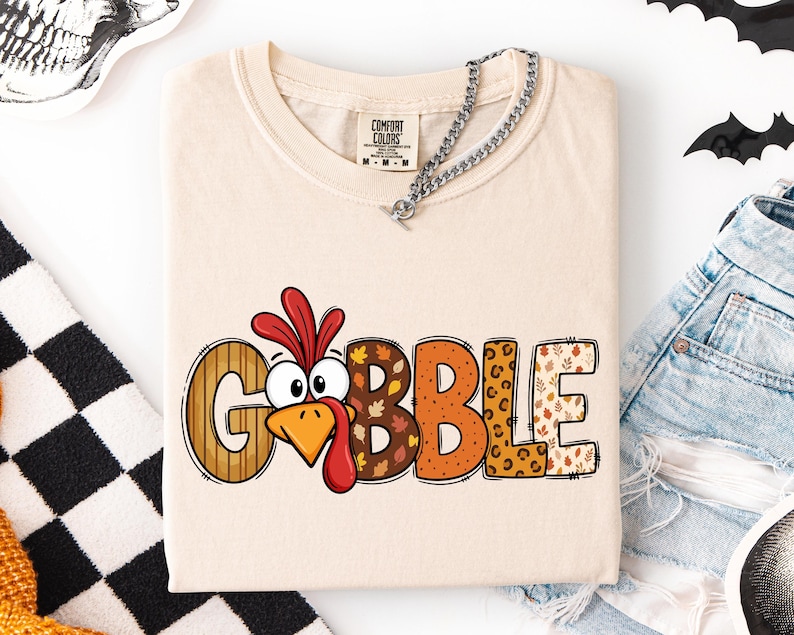 May include: A beige short-sleeved t-shirt featuring a graphic of a turkey head replacing the letter O in the word GOBBLE. The letters are filled with various patterns including stripes, autumn leaves, orange dots, and leopard print.