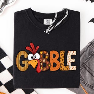 May include: A black long-sleeve shirt featuring the text GOBBLE in large, multi-patterned letters. The letter O is replaced by a cartoon turkey face with a red wattle and orange beak. The letters feature stripes, autumn leaves, and leopard print.