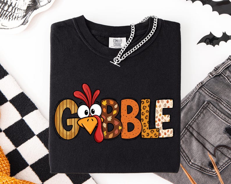 May include: A black long-sleeve shirt featuring the text GOBBLE in large, multi-patterned letters. The letter O is replaced by a cartoon turkey face with a red wattle and orange beak. The letters feature stripes, autumn leaves, and leopard print.