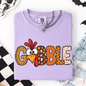 May include: A light purple short-sleeve t-shirt featuring the word GOBBLE in large, multi-patterned letters. The letter O is replaced by a cartoon turkey face with a red wattle and orange beak. The other letters feature stripes, autumn leaves, and leopard print.