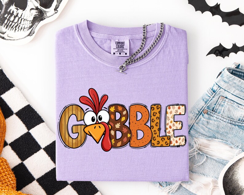 May include: A light purple short-sleeve t-shirt featuring the word GOBBLE in large, multi-patterned letters. The letter O is replaced by a cartoon turkey face with a red wattle and orange beak. The other letters feature stripes, autumn leaves, and leopard print.