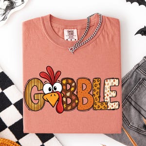 May include: A dusty rose t-shirt featuring the word GOBBLE in large, patterned letters. The letter O is replaced by a cartoon turkey head with a red wattle and orange beak. The other letters feature stripes, autumn leaves, and leopard print.
