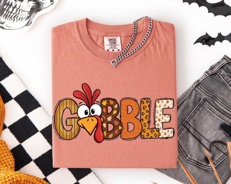 May include: A dusty rose t-shirt featuring the word GOBBLE in large, patterned letters. The letter O is replaced by a cartoon turkey head with a red wattle and orange beak. The other letters feature stripes, autumn leaves, and leopard print.