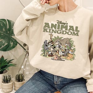 May include: A beige sweatshirt with the text "Disney's Animal Kingdom" in green above a colourful graphic of cartoon animals. The graphic includes a zebra, giraffe, elephant, and other characters.