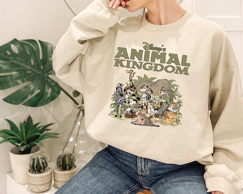 May include: A beige sweatshirt with the text "Disney's Animal Kingdom" in green above a colourful graphic of cartoon animals. The graphic includes a zebra, giraffe, elephant, and other characters.