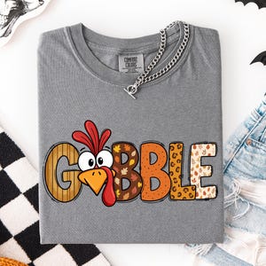 May include: A grey short-sleeved t-shirt featuring the word GOBBLE in large, multi-patterned letters. The letter O is replaced by a cartoon turkey face with a red wattle and orange beak. The other letters feature wood grain, leaf, and leopard prints.
