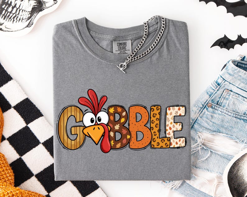 May include: A grey short-sleeved t-shirt featuring the word GOBBLE in large, multi-patterned letters. The letter O is replaced by a cartoon turkey face with a red wattle and orange beak. The other letters feature wood grain, leaf, and leopard prints.