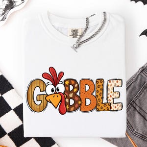 May include: A white t-shirt featuring the word GOBBLE printed in large, colorful letters. The letter O is replaced by a cartoon turkey face with a red wattle. The letters feature various patterns including wood grain, autumn leaves, and leopard print.
