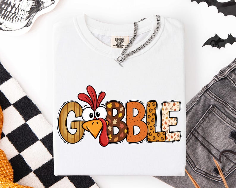 May include: A white t-shirt featuring the word GOBBLE printed in large, colorful letters. The letter O is replaced by a cartoon turkey face with a red wattle. The letters feature various patterns including wood grain, autumn leaves, and leopard print.